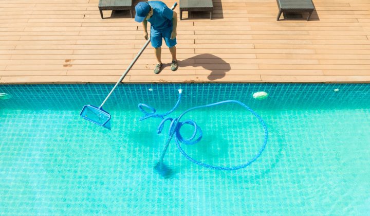 A technician in blue uniform cleans a large outdoor pool with a vacuum hose and skimmer net, viewed from above on a sunny day with wooden decking and lounge chairs nearby.