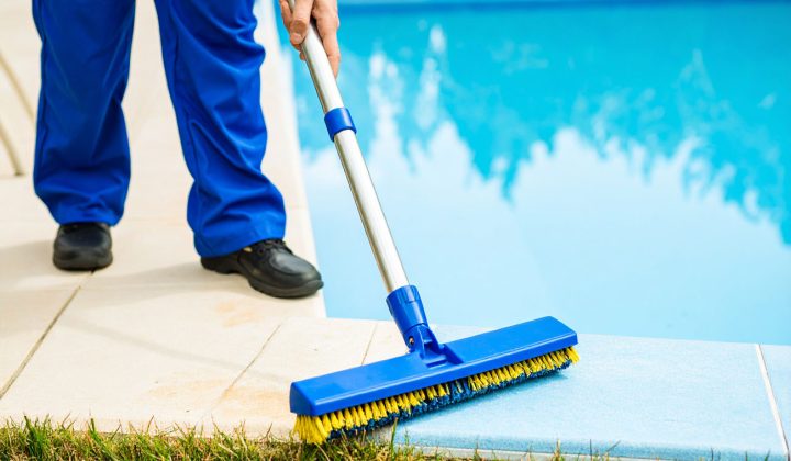 A worker in blue overalls uses a long-handled scrub brush to clean the tiled edge of a swimming pool deck.