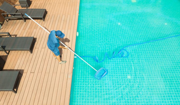 A worker in blue attire uses a long-handled skimmer net to clean the surface of a large outdoor pool surrounded by wooden decking and lounge chairs.
