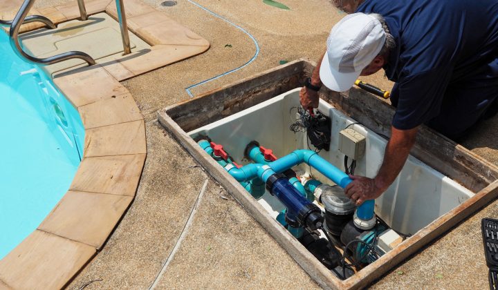 A technician working on the pool's filtration system, adjusting valves and components within an open access panel near the pool edge.