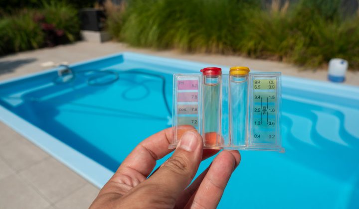 A hand holding a pool water test kit displaying pH and chlorine levels, with a clean blue pool in the background.