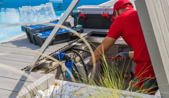 A technician in a red uniform works on pool plumbing components near a maintenance access point, with tools and equipment organized nearby.