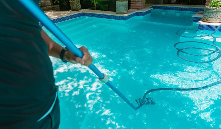 A person using a pool vacuum attachment to clean the bottom of a clear, turquoise swimming pool with sunlight reflecting on the surface.